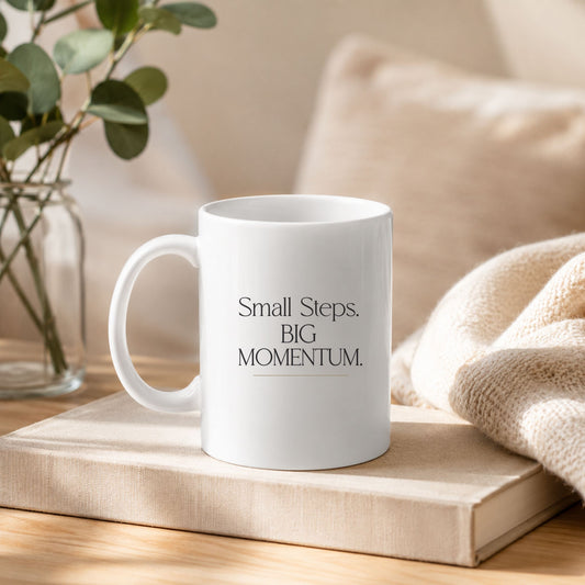 White ceramic mug with the Small Steps. BIG MOMENTUM. design displayed on a book in a cozy neutral-toned home setting with soft textures and warm natural light.