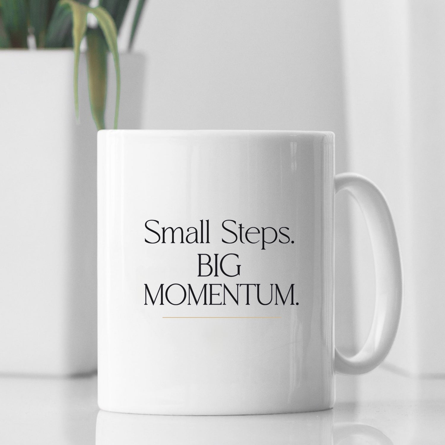 White ceramic mug with "Small Steps. BIG Momentum." design on a minimal table with soft natural light and plant background