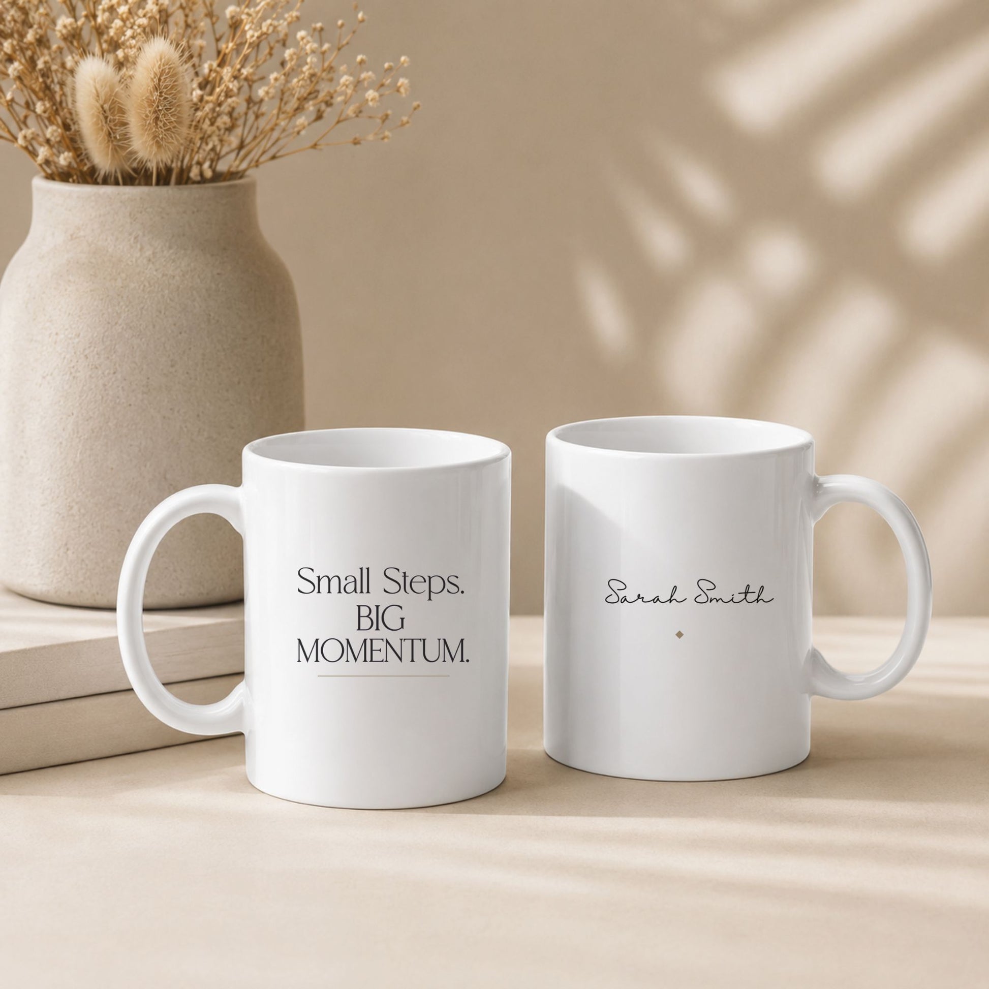 Two white ceramic mugs featuring the Small Steps. BIG MOMENTUM. design and personalized name option, displayed in a cozy neutral-toned lifestyle setting. Momentum Prints.
