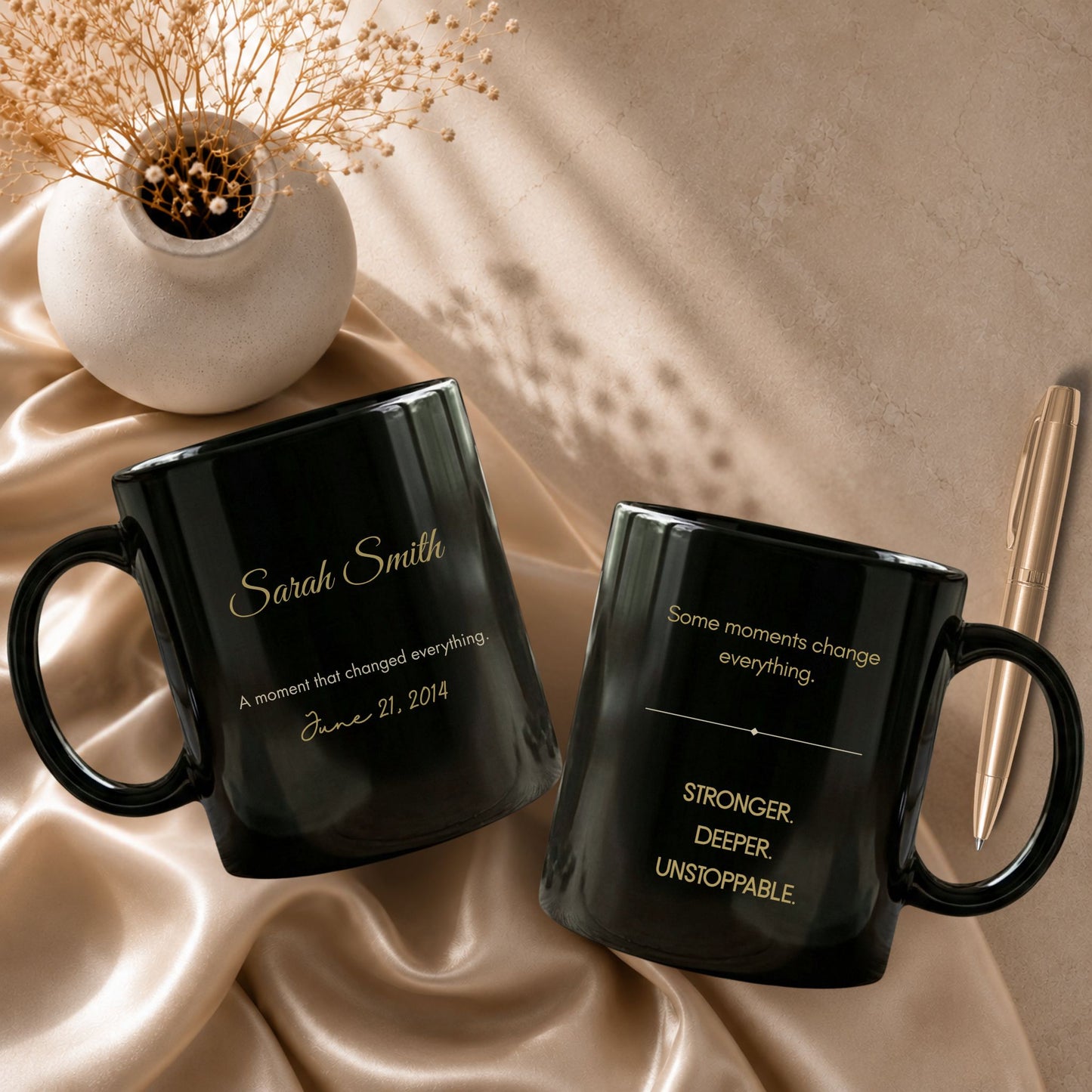 Two personalized black ceramic mugs displayed on champagne satin fabric with dried baby's breath and a gold pen, showcasing the name side and the affirmation side by Momentum Prints.