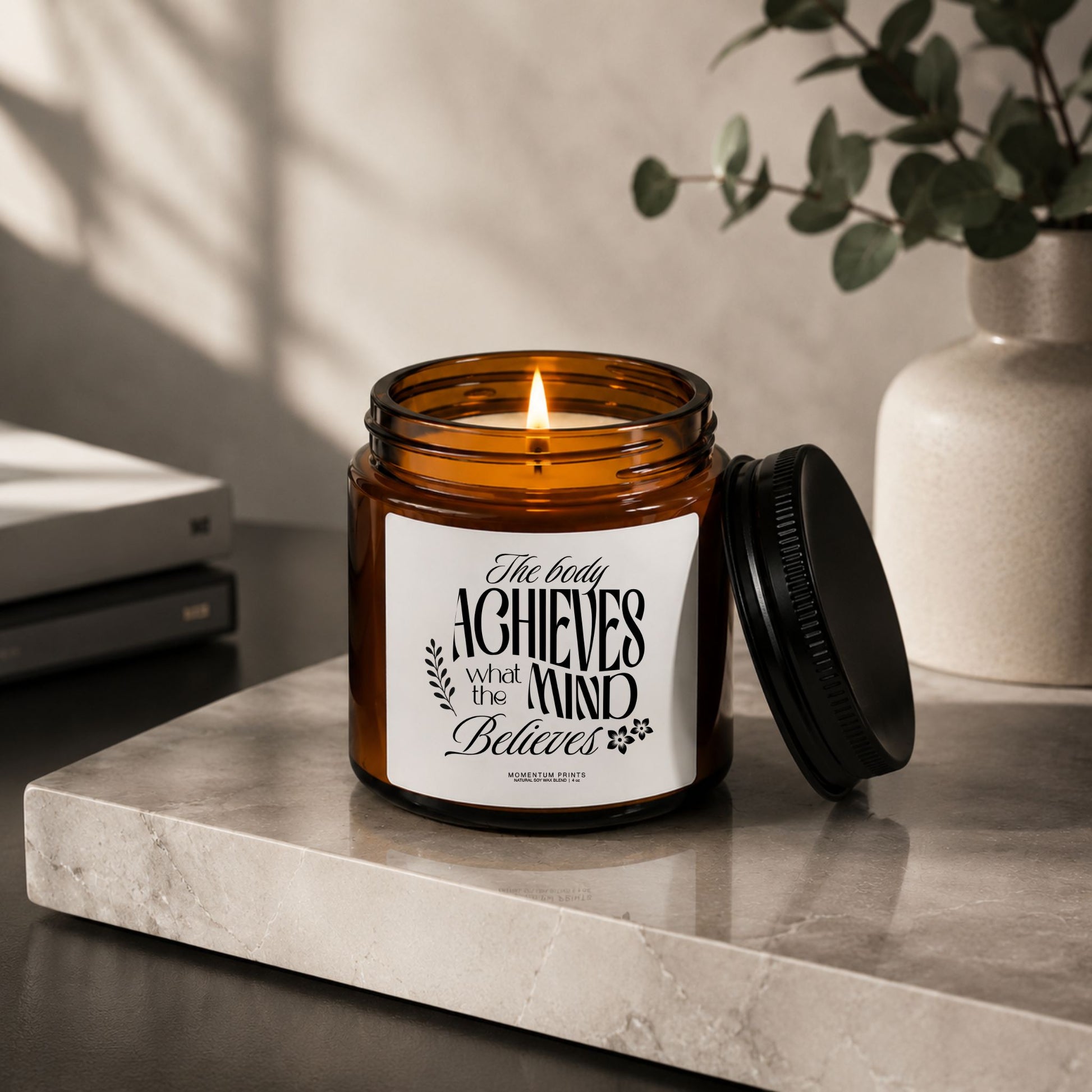 The Body Achieves What the Mind Believes 4 oz amber glass candle with black lid beside the jar on a marble surface with modern neutral decor.
