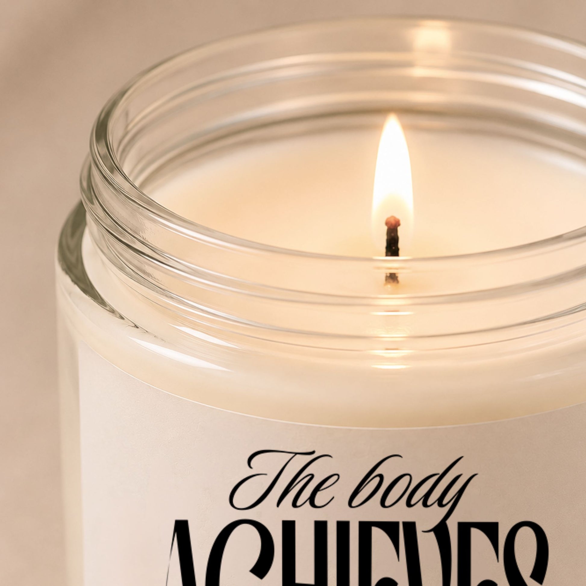 The Body Achieves What the Mind Believes 9 oz clear glass soy candle close-up featuring a soft glowing flame, smooth white wax, and minimalist neutral styling.