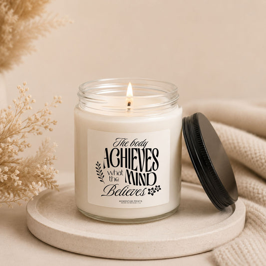 The Body Achieves What the Mind Believes 9 oz clear glass candle with black lid beside the jar, styled with soft beige decor and cozy neutral textures.