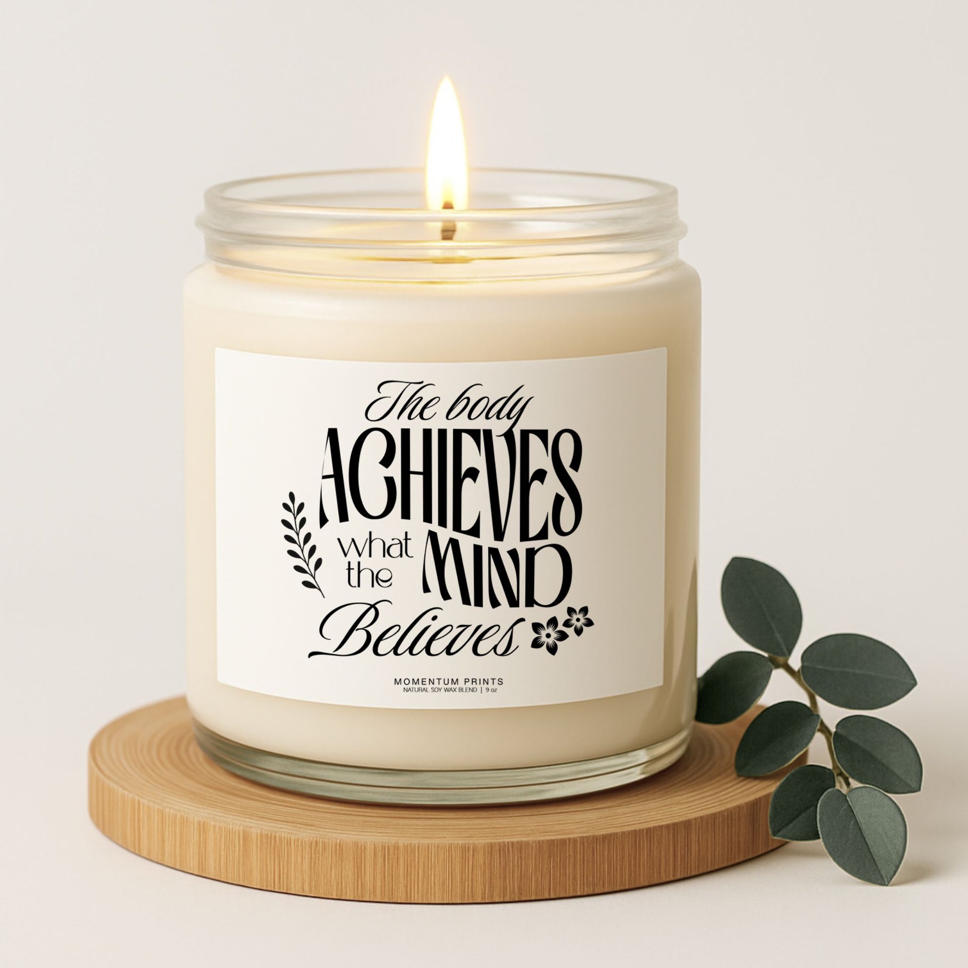 The Body Achieves What the Mind Believes 9 oz clear glass candle with eucalyptus leaves and soft natural lighting in a clean minimalist setting.