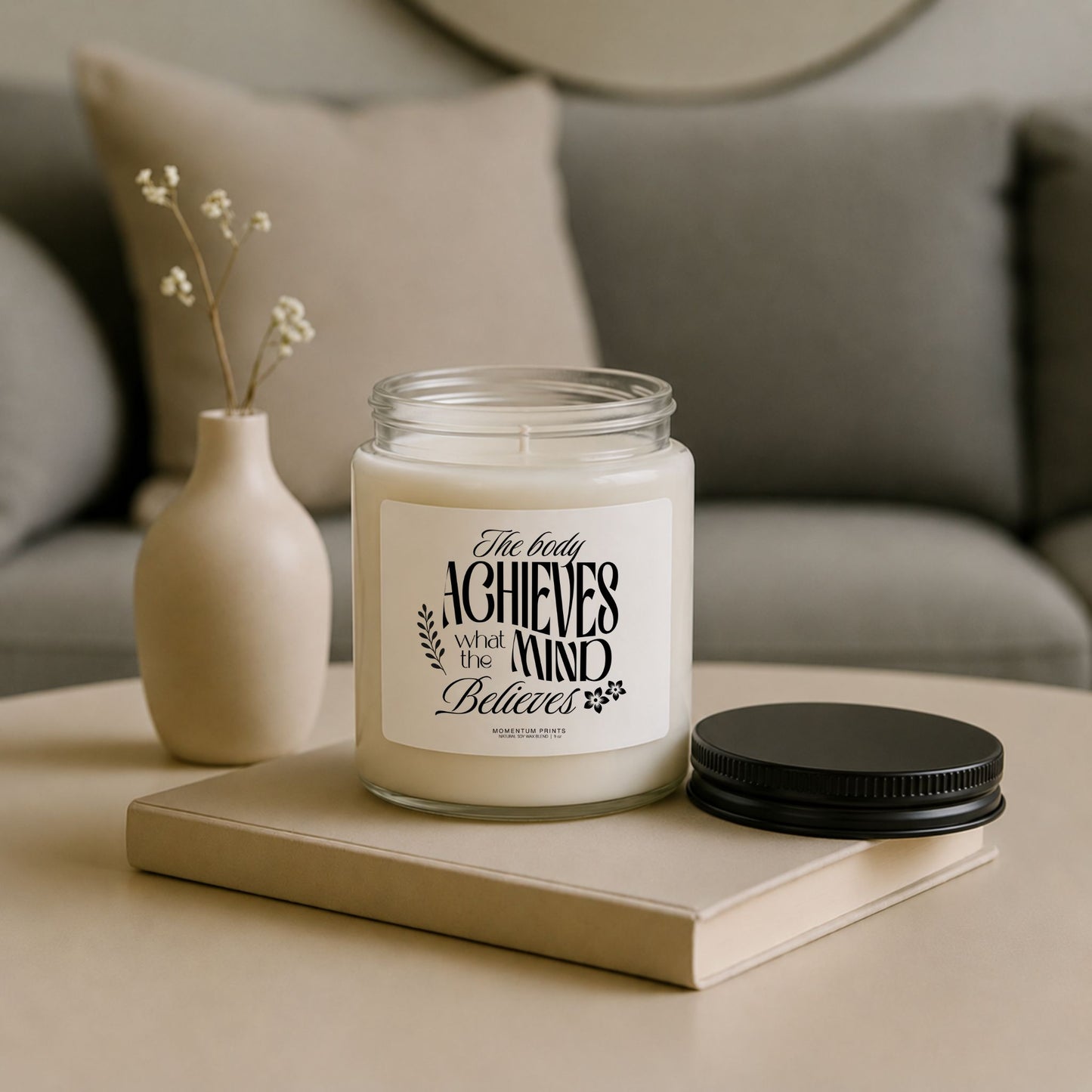 The Body Achieves What the Mind Believes 9 oz clear glass soy candle styled on a neutral coffee table with minimalist decor and black lid beside the jar.