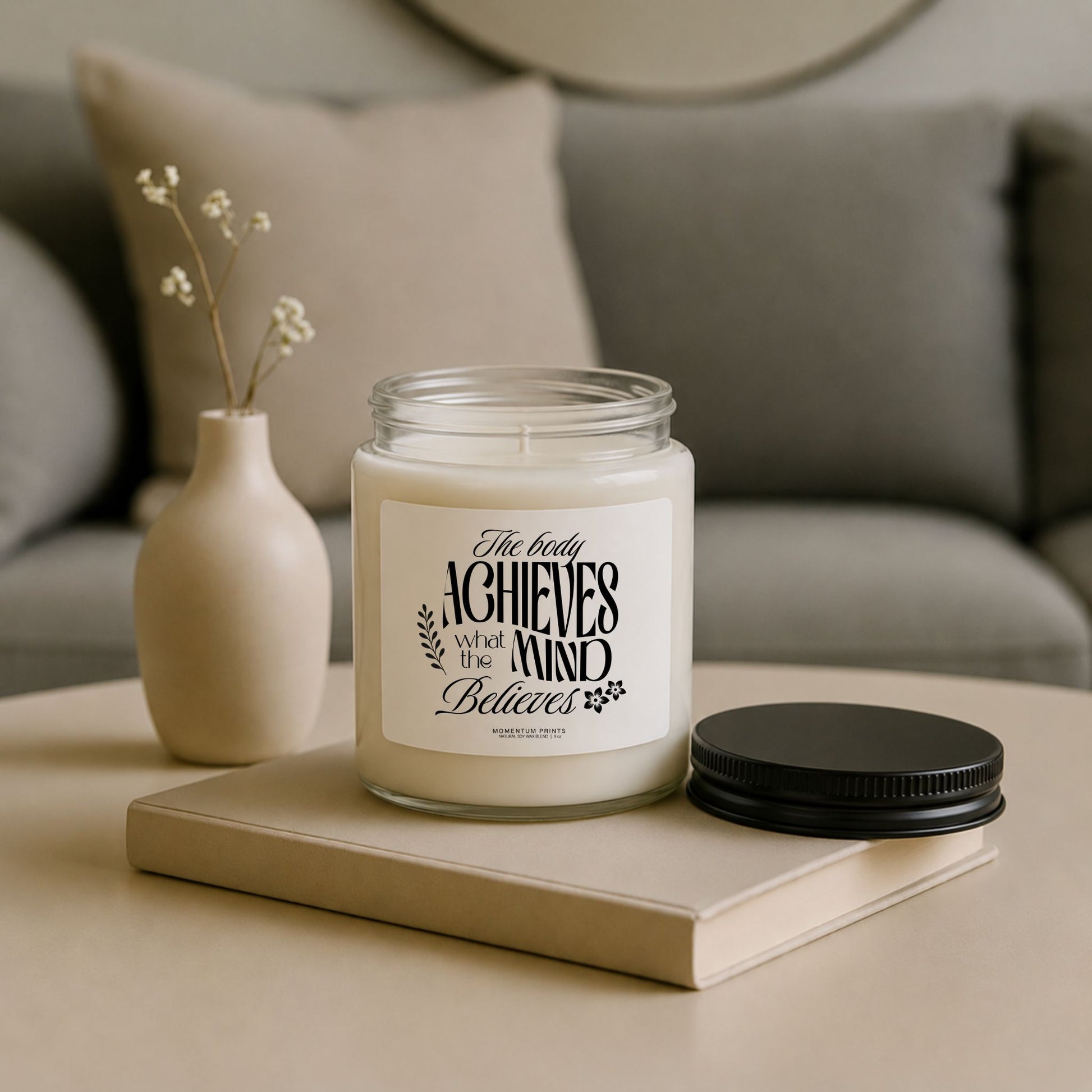 The Body Achieves What the Mind Believes 9 oz clear glass soy candle styled on a neutral coffee table with minimalist decor and black lid beside the jar.