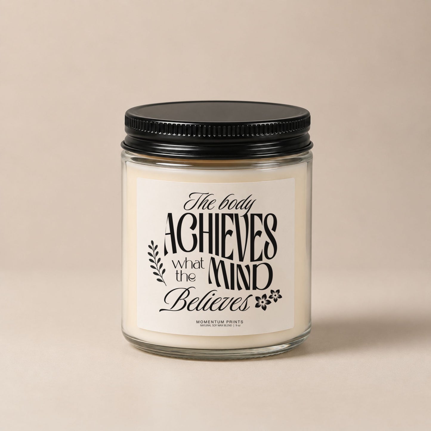 The Body Achieves What the Mind Believes 9 oz clear glass candle with black lid against a soft beige minimalist background.