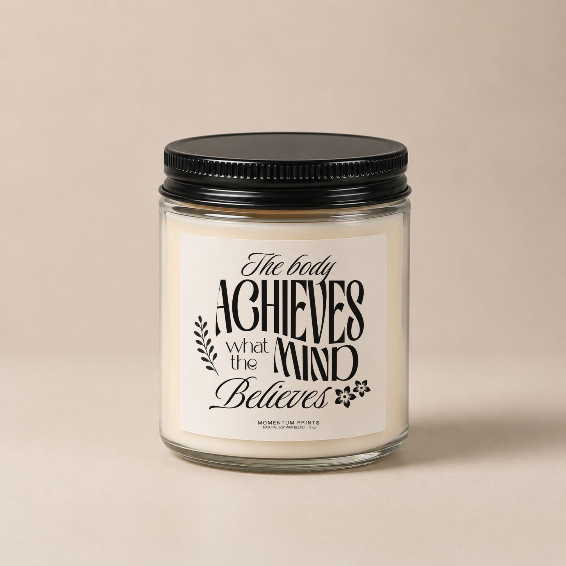 The Body Achieves What the Mind Believes 9 oz clear glass candle with black lid against a soft beige minimalist background.