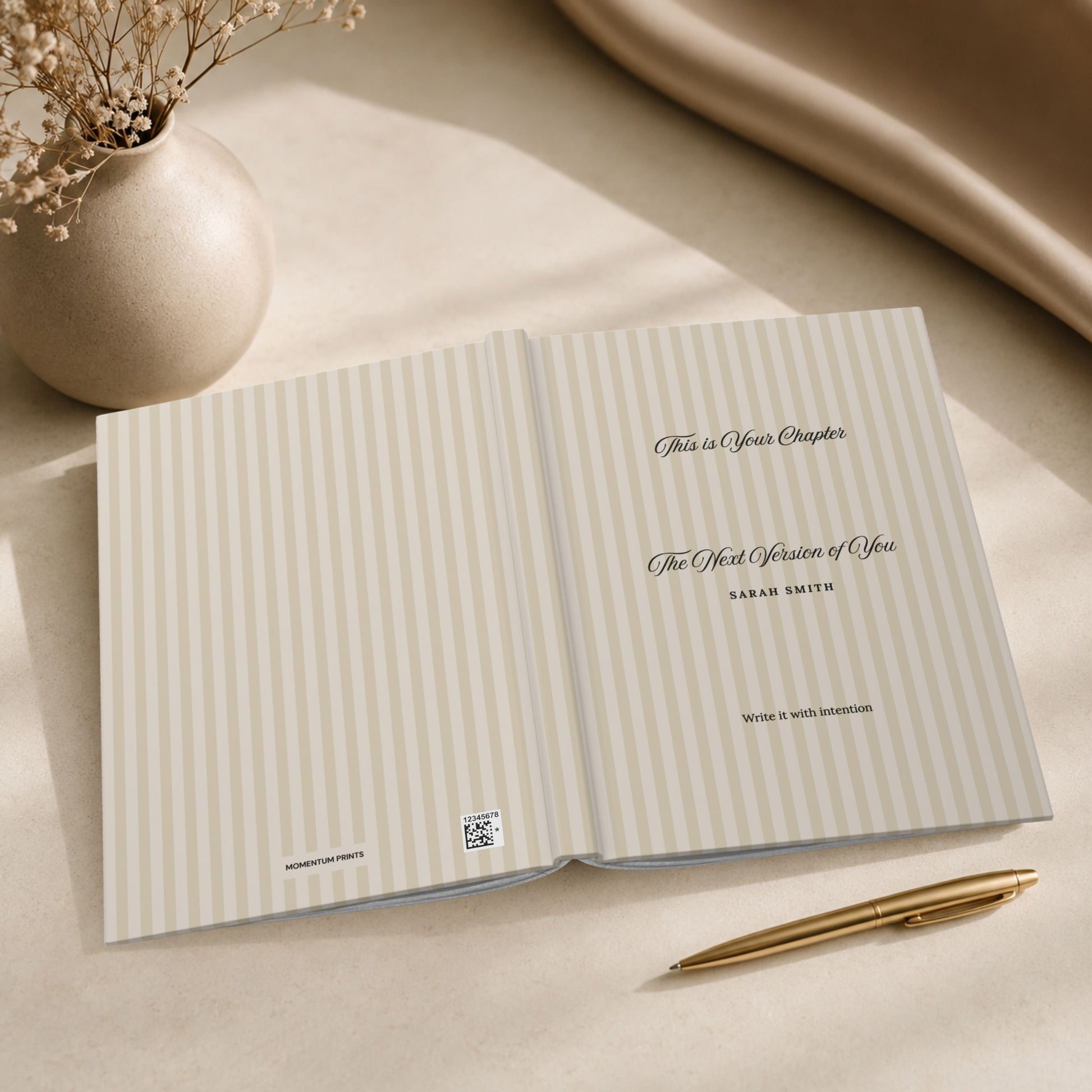 Open personalized hardcover journal with beige striped cover displaying the interior title page in a minimalist editorial lifestyle scene with ceramic vase and gold pen by Momentum Prints.