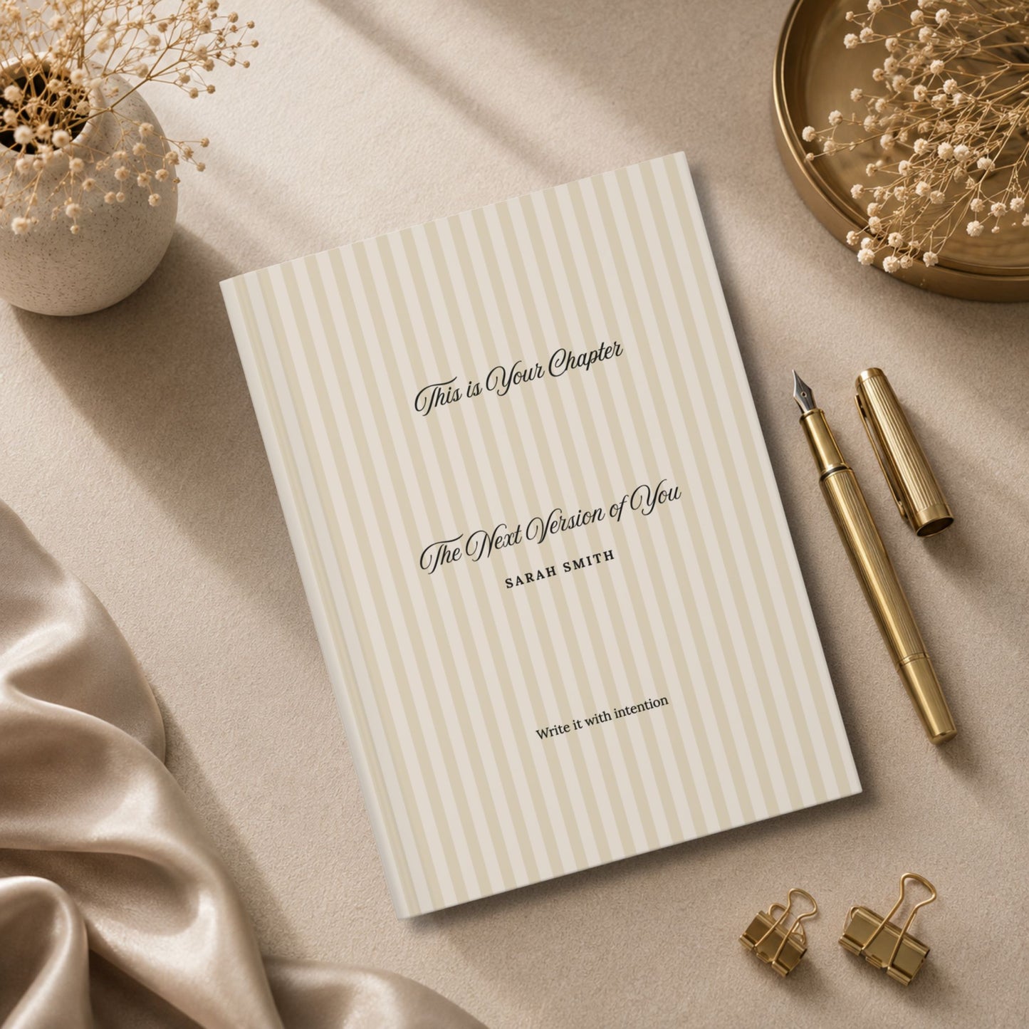 Premium personalized hardcover journal with beige striped cover standing beside champagne gold pen, matte ceramic vase and beige silk fabric in a luxury editorial lifestyle setting by Momentum Prints