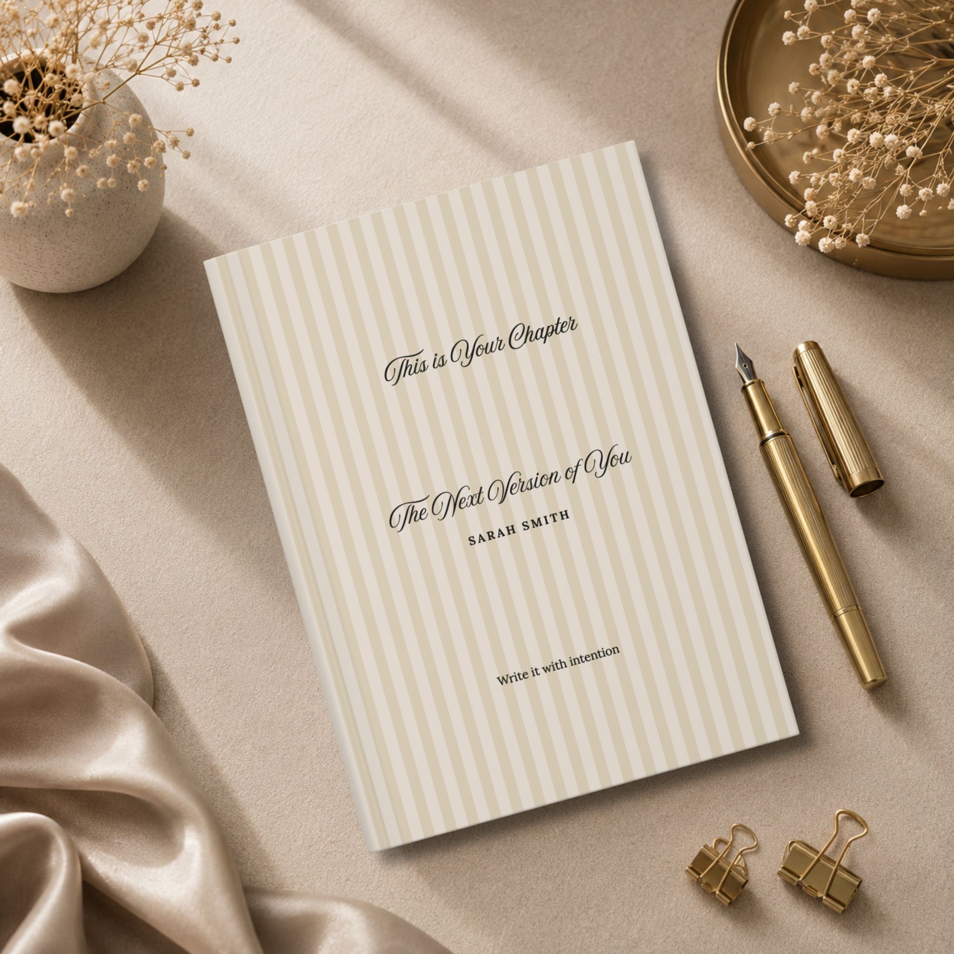 Premium personalized hardcover journal with beige striped cover standing beside champagne gold pen, matte ceramic vase and beige silk fabric in a luxury editorial lifestyle setting by Momentum Prints