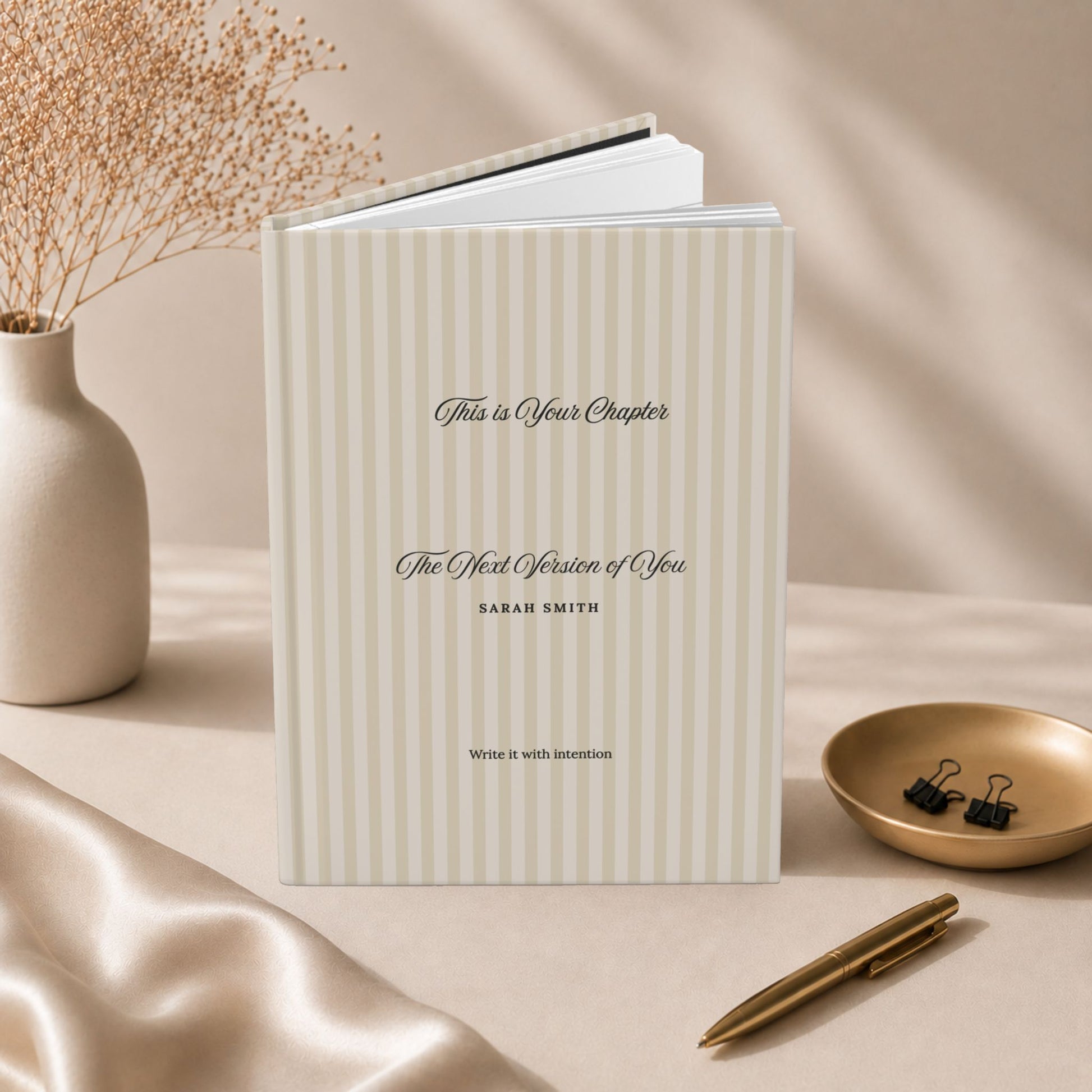 Premium personalized hardcover journal with beige striped cover standing beside champagne gold pen, matte ceramic vase and beige silk fabric in a luxury editorial lifestyle setting by Momentum Prints.