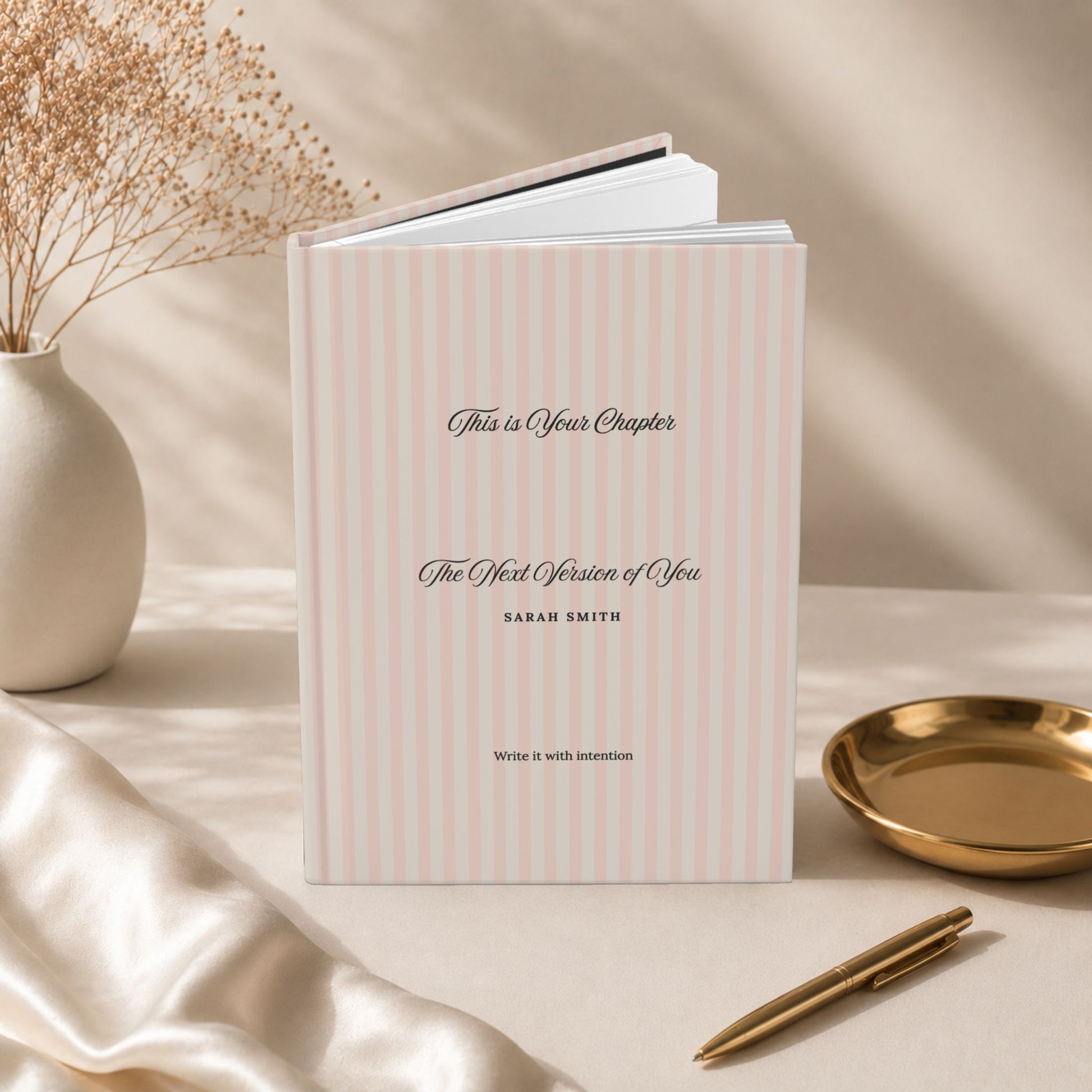 Premium personalized hardcover journal with dusty rose striped cover standing upright with lined pages fanned open, styled beside dried flowers in a ceramic vase and a small gold dish in a warm editorial setting by Momentum Prints.