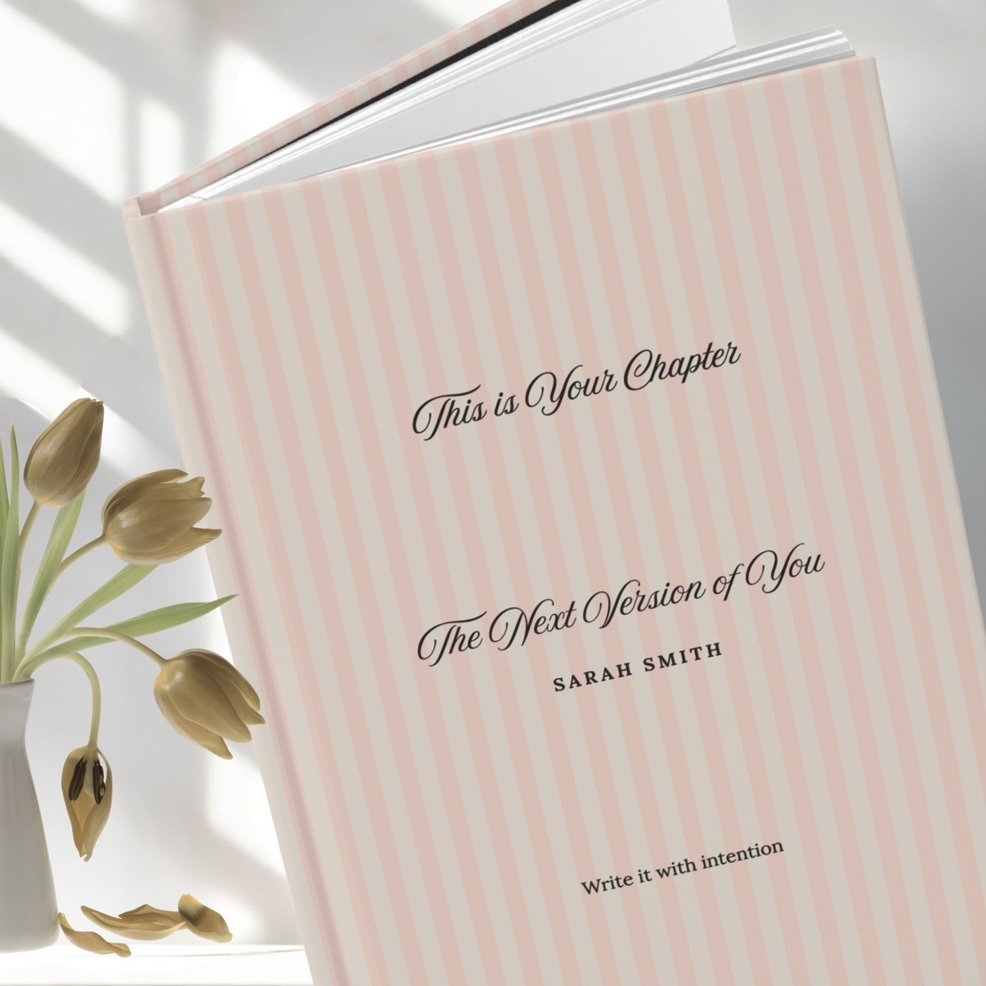 Premium personalized hardcover journal with dusty rose striped cover standing upright beside dried gold tulips in a white ceramic vase, set against a bright airy backdrop by Momentum Prints.