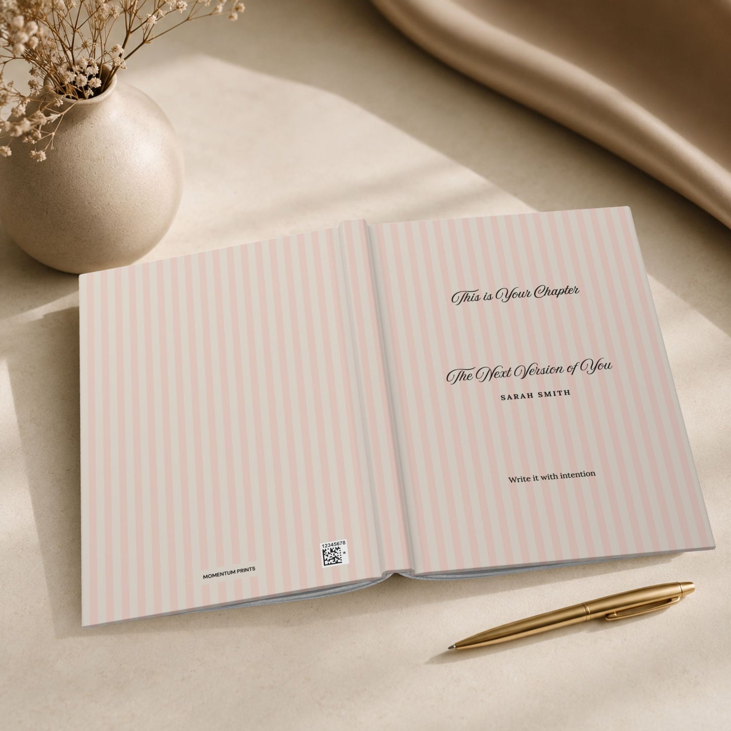 Premium personalized hardcover journal with dusty rose striped cover shown open flat, revealing the inside cover and title page, styled beside a ceramic vase with dried flowers and draped fabric in a warm editorial setting by Momentum Prints