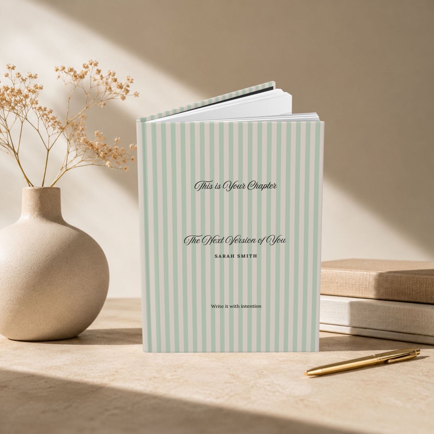 Premium personalized hardcover journal with sage green striped cover standing upright on a stone desk beside a ceramic vase with dried flowers and a stack of books, in a warm sunlit lifestyle setting by Momentum Prints.