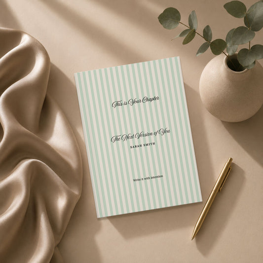 Premium personalized hardcover journal with sage green striped cover styled in a flat lay with a gold pen, draped silk fabric, and a ceramic vase with eucalyptus in a warm editorial setting by Momentum Prints.