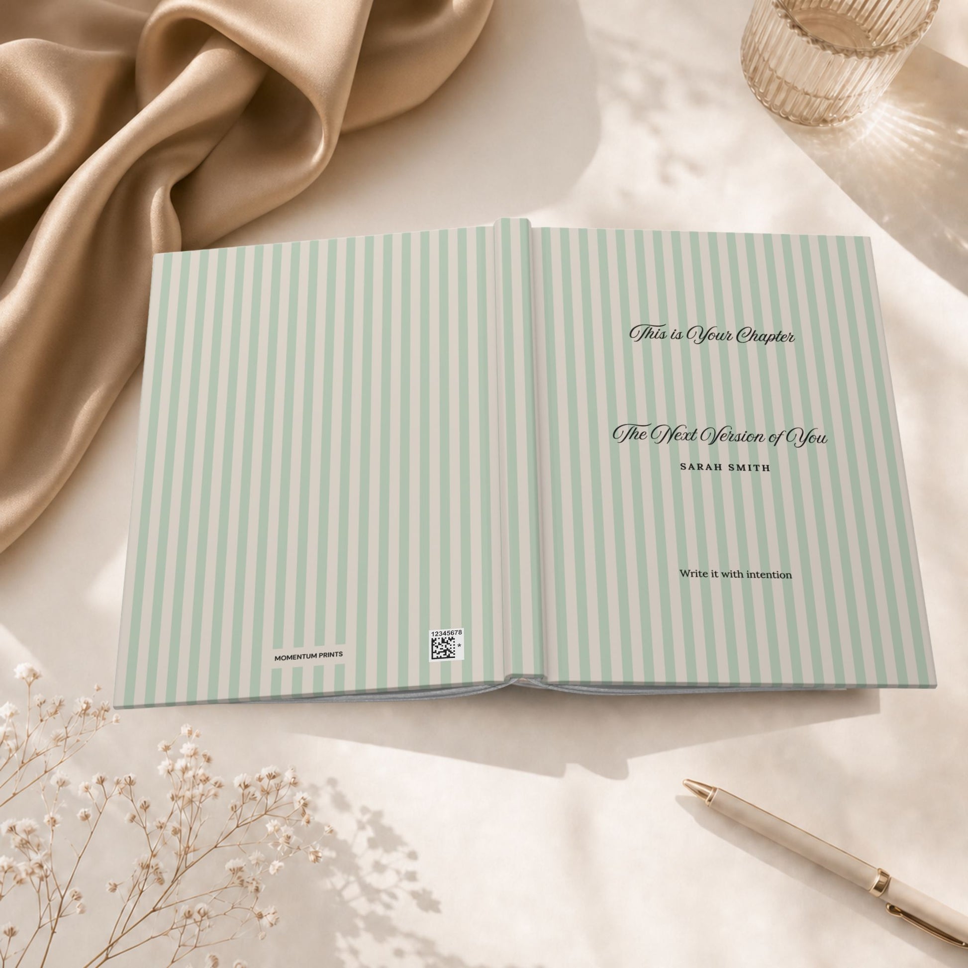 Premium personalized hardcover journal with sage green striped cover shown open flat, revealing the inside cover and title page, styled beside draped silk fabric and a glass candle holder in a warm editorial setting by Momentum Prints.