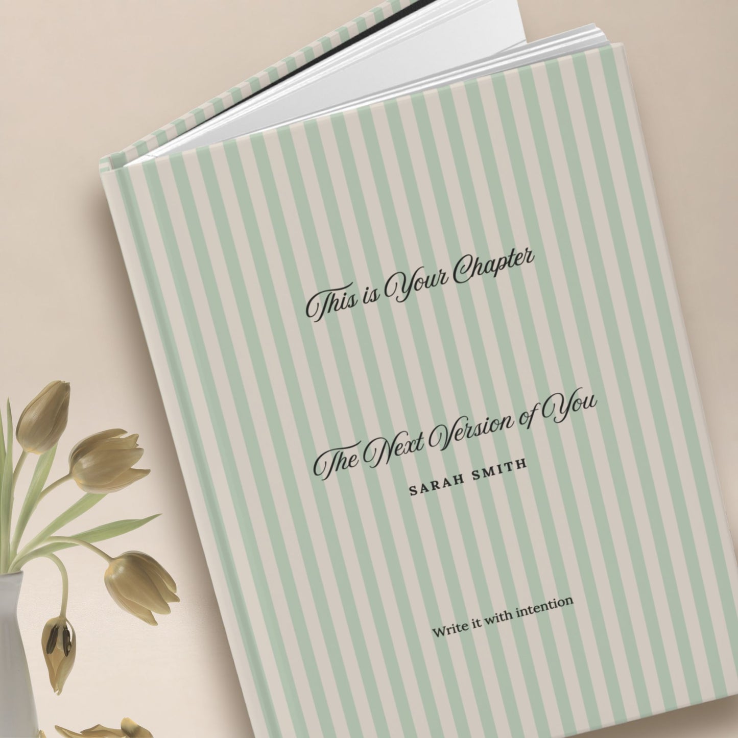 Premium personalized hardcover journal with sage green striped cover standing upright beside dried gold tulips in a vase, set against a warm neutral backdrop by Momentum Prints.