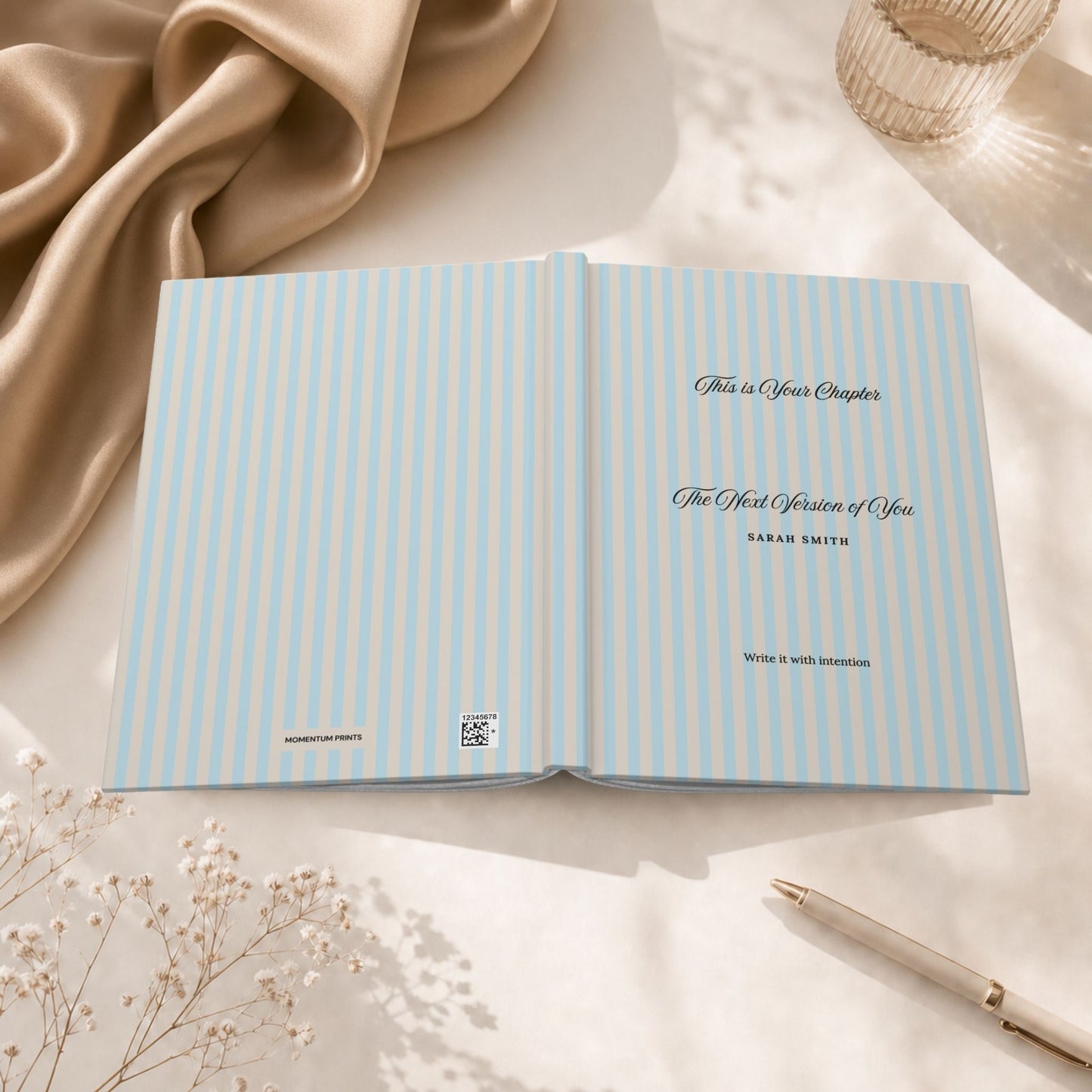 Premium personalized hardcover journal with sky blue striped cover shown open flat, revealing the inside cover and title page, styled beside a glass candle holder and draped silk fabric in a warm editorial setting by Momentum Prints.