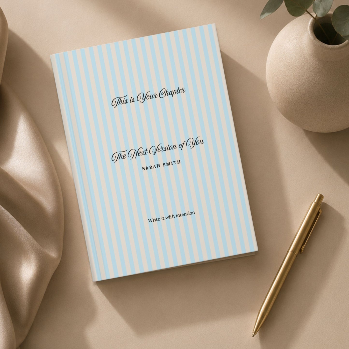 Premium personalized hardcover journal with sky blue striped cover styled in a flat lay beside draped silk fabric, a ceramic vase with eucalyptus, and a gold pen in a luxury editorial setting by Momentum Prints.