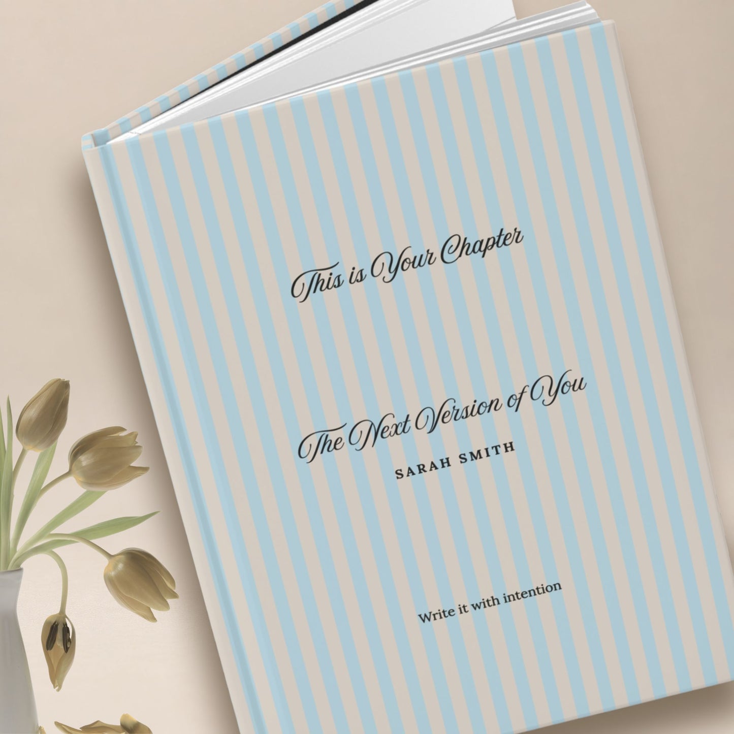 Premium personalized hardcover journal with sky blue striped cover standing upright beside dried gold tulips in a vase, set against a warm neutral backdrop by Momentum Prints.