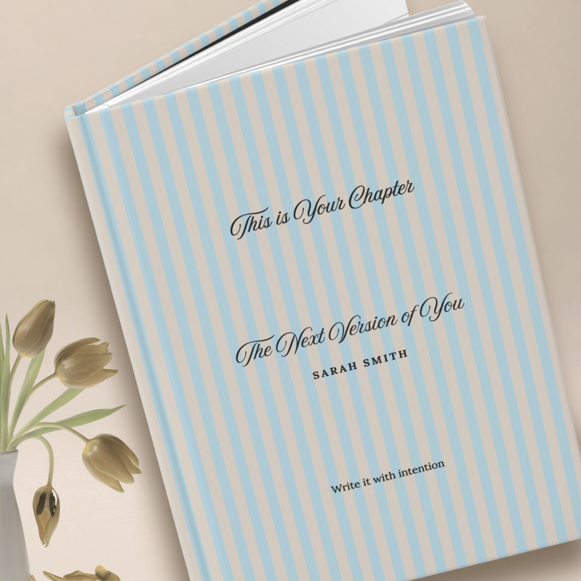 Premium personalized hardcover journal with sky blue striped cover standing upright beside dried gold tulips in a vase, set against a warm neutral backdrop by Momentum Prints.