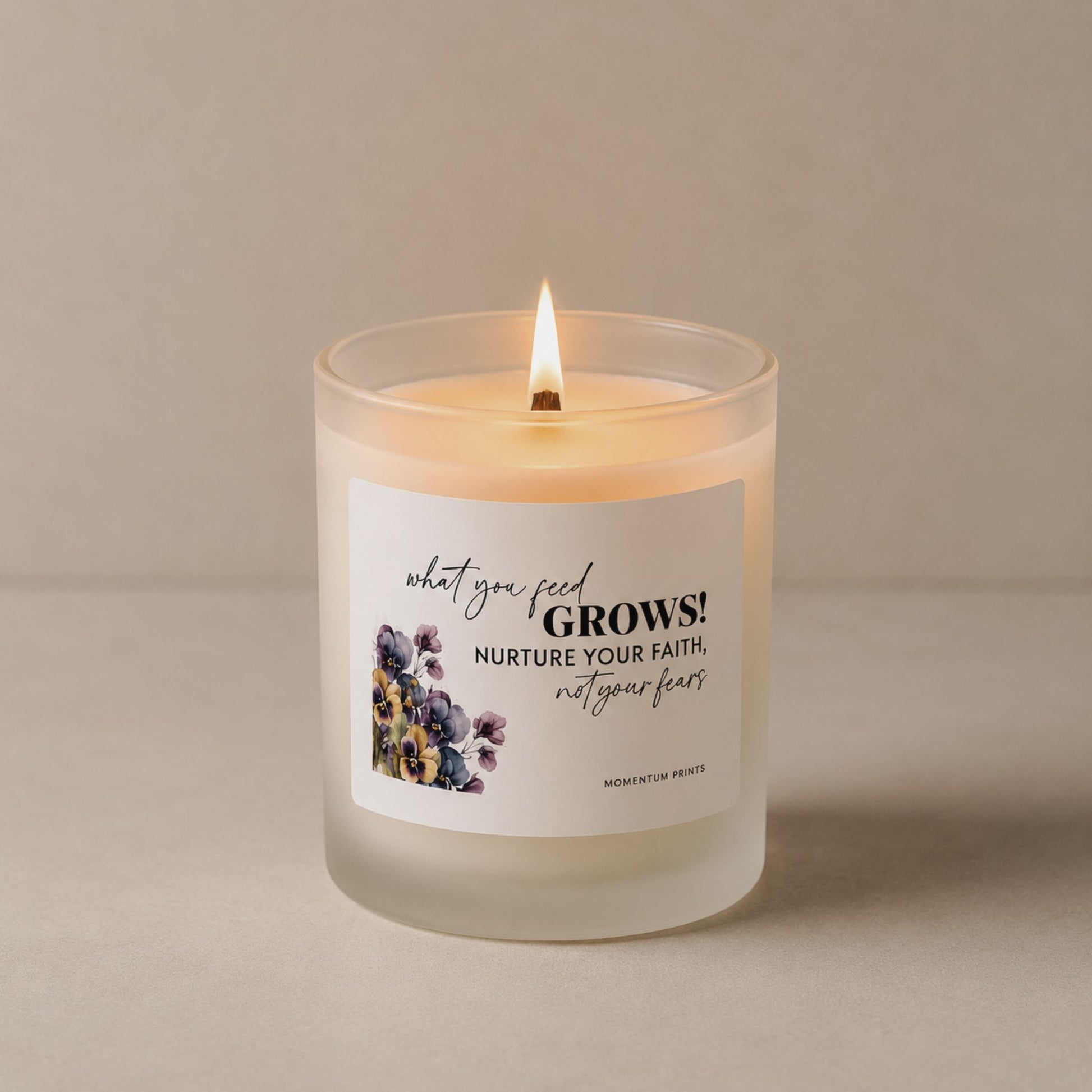 Lit frosted glass candle with the phrase “What You Feed Grows” by Momentum Prints, styled in a minimalist neutral setting with soft warm lighting and a wooden wick flame.