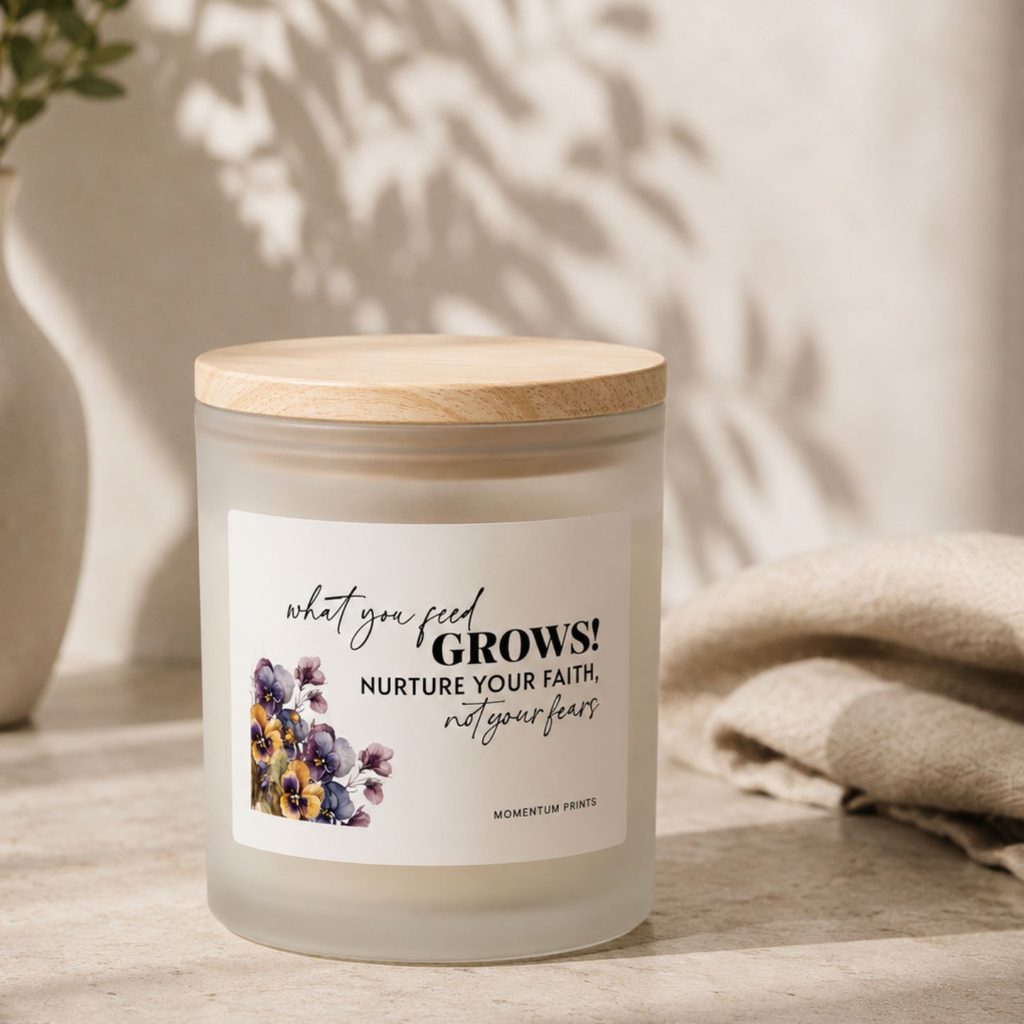 Frosted glass candle with “What You Feed Grows” label by Momentum Prints, styled in a minimalist neutral setting with soft shadows, natural textures, and warm ambient lighting.